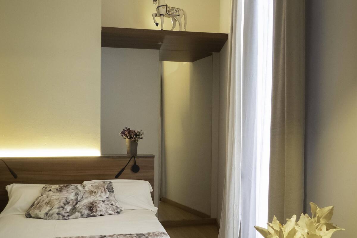 Quarto de hotel com cama, mesa de cabeceira com um vaso de flores, uma cadeira e uma pequena mesa.