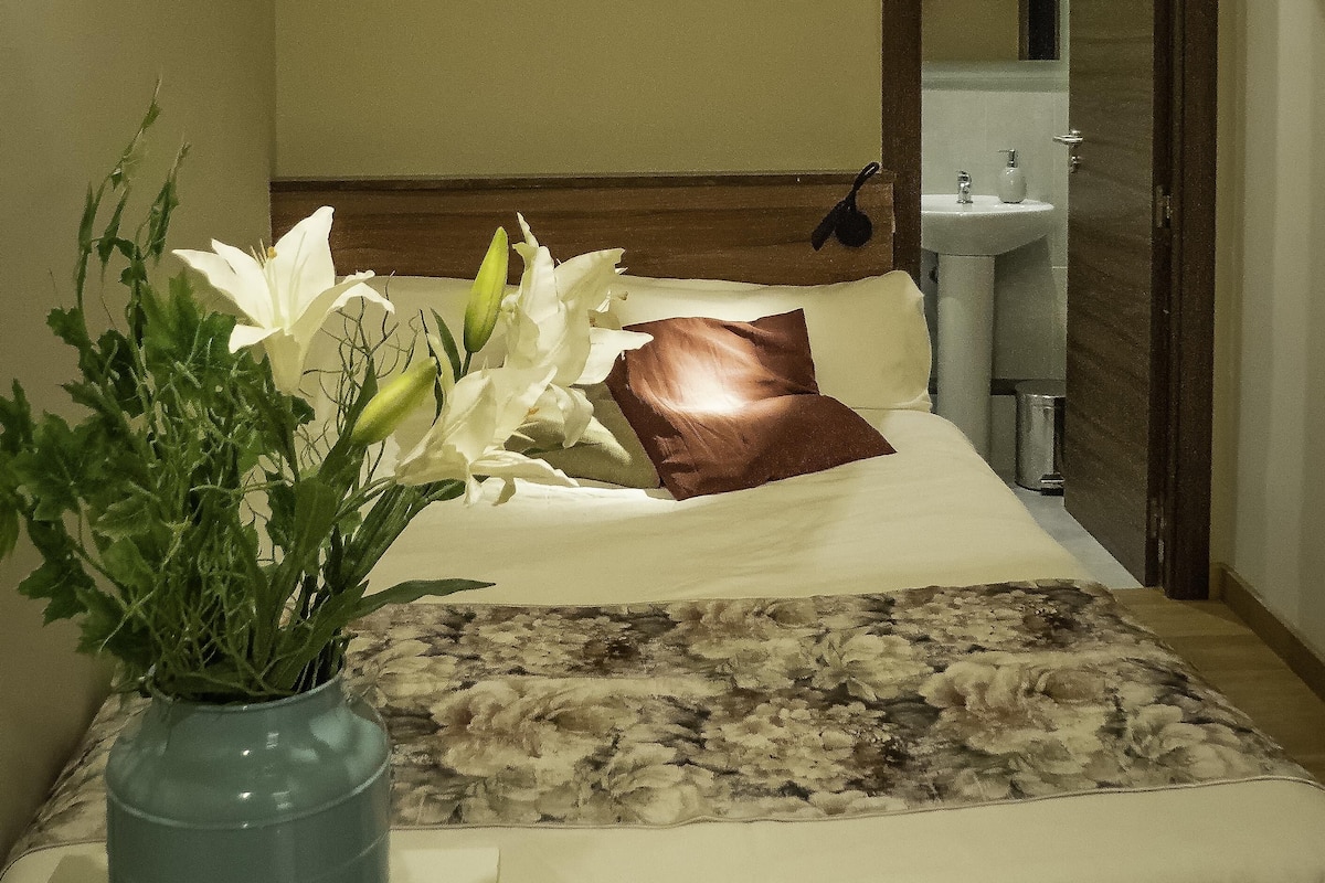 Quarto de hotel com uma cama, um vaso de flores, um banheiro visível através de uma porta aberta e um ar-condicionado fixado na parede.