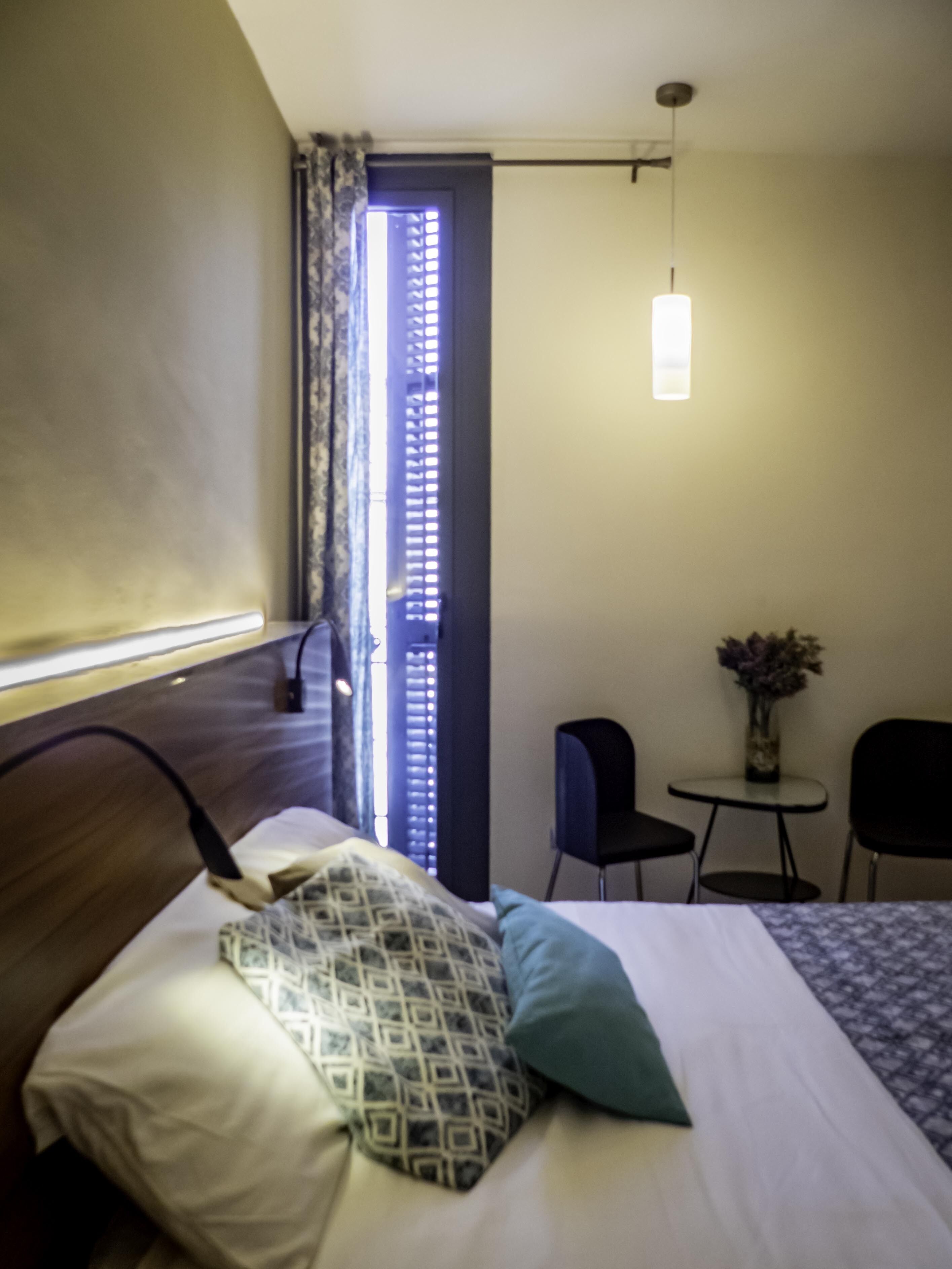Hotelkamer met een bed, bureau en stoel. Er is een raam met gordijnen, een hanglamp en een klein tafeltje met een bloevas.