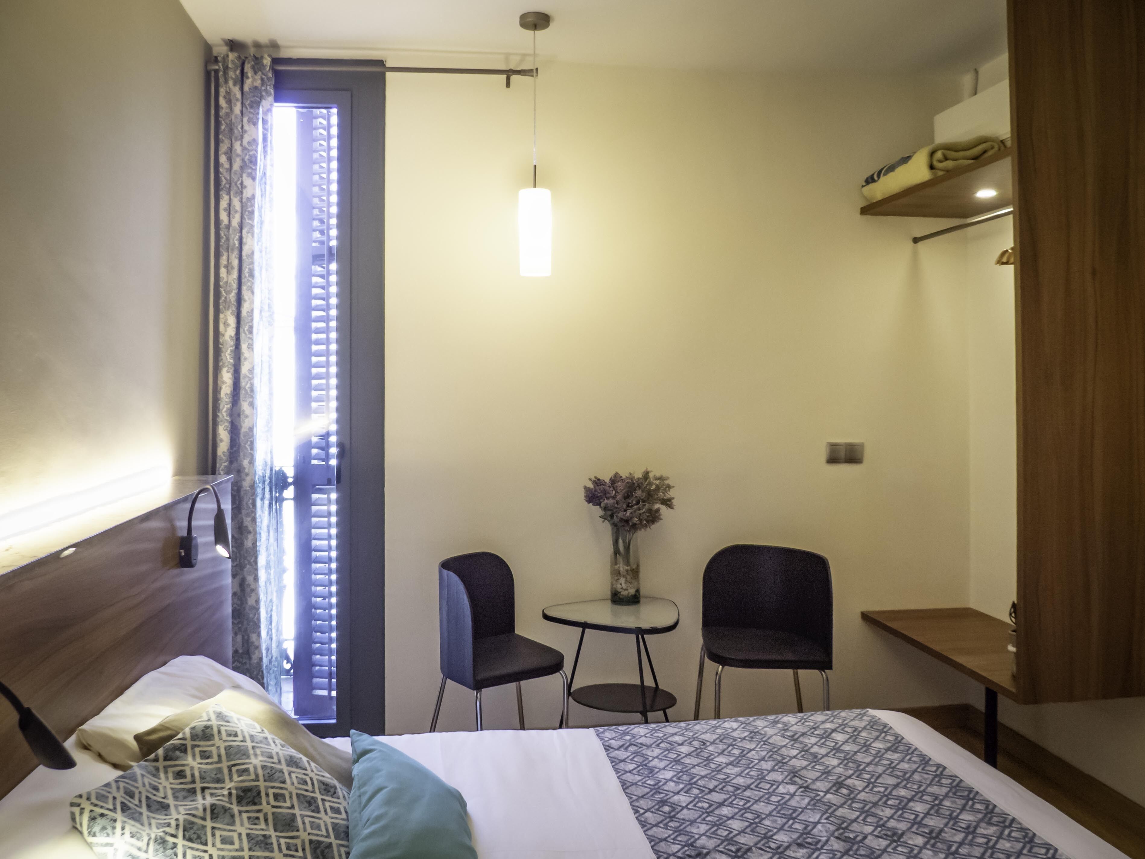 A hotel room with a bed, a small table, two chairs, a vase with flowers, and a shelf with folded towels.