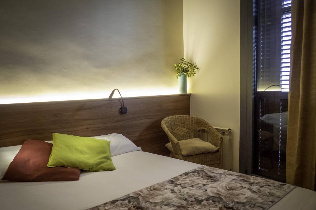 Quarto de hotel com cama, uma cadeira, luminária na parede e janela com persianas.