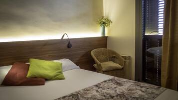 Una habitación de hotel con cama, una silla, una lámpara de pared y una ventana con persianas.