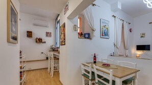 A dining area with a wooden table and white chairs, a small kitchen area with a stove and shelves, and a TV mounted on the wall. - Gladiator's House (Rome)