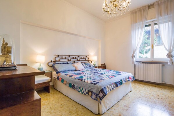 A bedroom with a bed, bedside tables, a chandelier, a window with curtains, and a radiator. - Residenza Novecento (Verona)