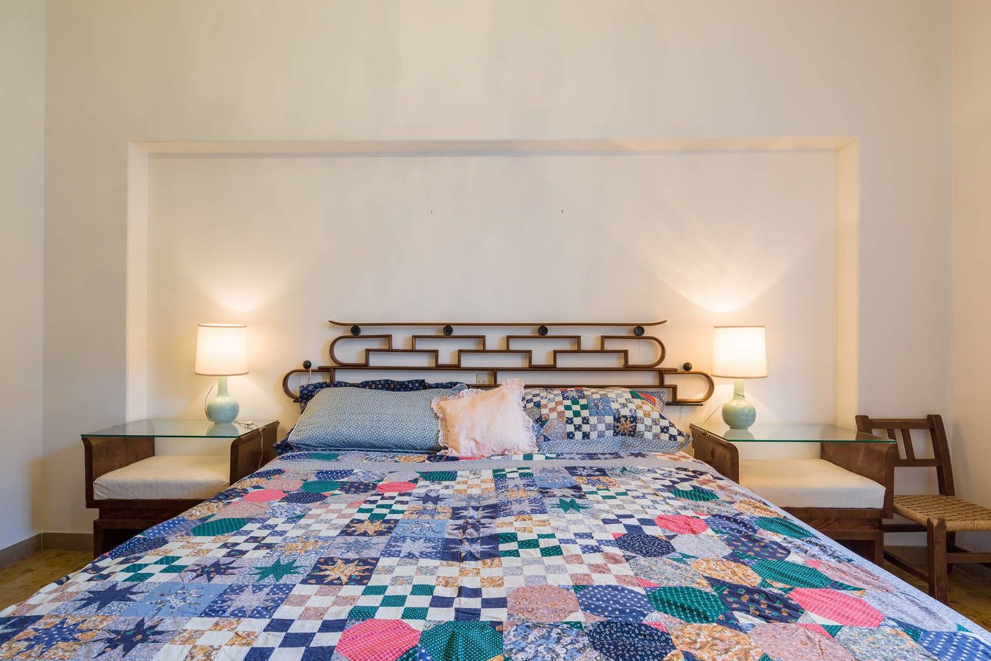 A neatly made bed with a colorful quilt, two bedside lamps, and a wooden nightstand with a chair.