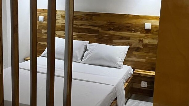 A bedroom with a wooden headboard, two bedside tables, a bed with white bedding, and two black wall-mounted lamps. - Résidence Mbemba (Abidjan)
