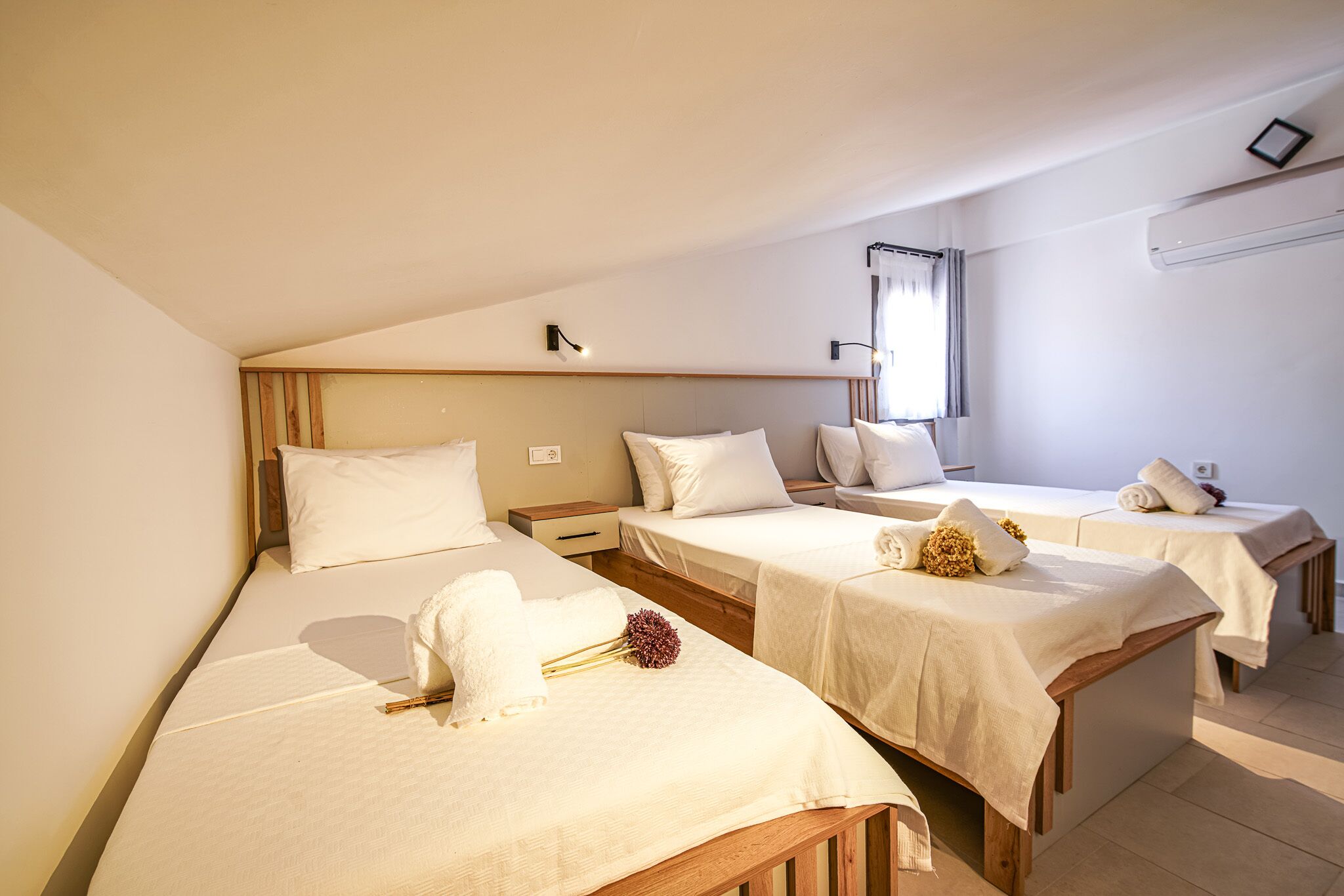 Habitación de hotel con tres camas individuales, cada una con colcha y almohadas blancas. Cada cama tiene una mesita de noche pequeña, y al fondo, una ventana con cortinas.