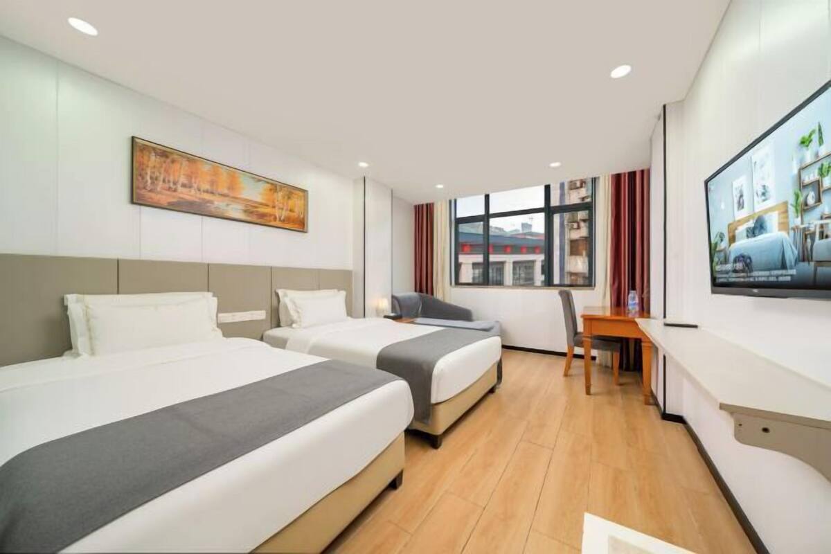 A hotel room with two beds, a desk, a chair, a TV, and a window with a view of buildings.