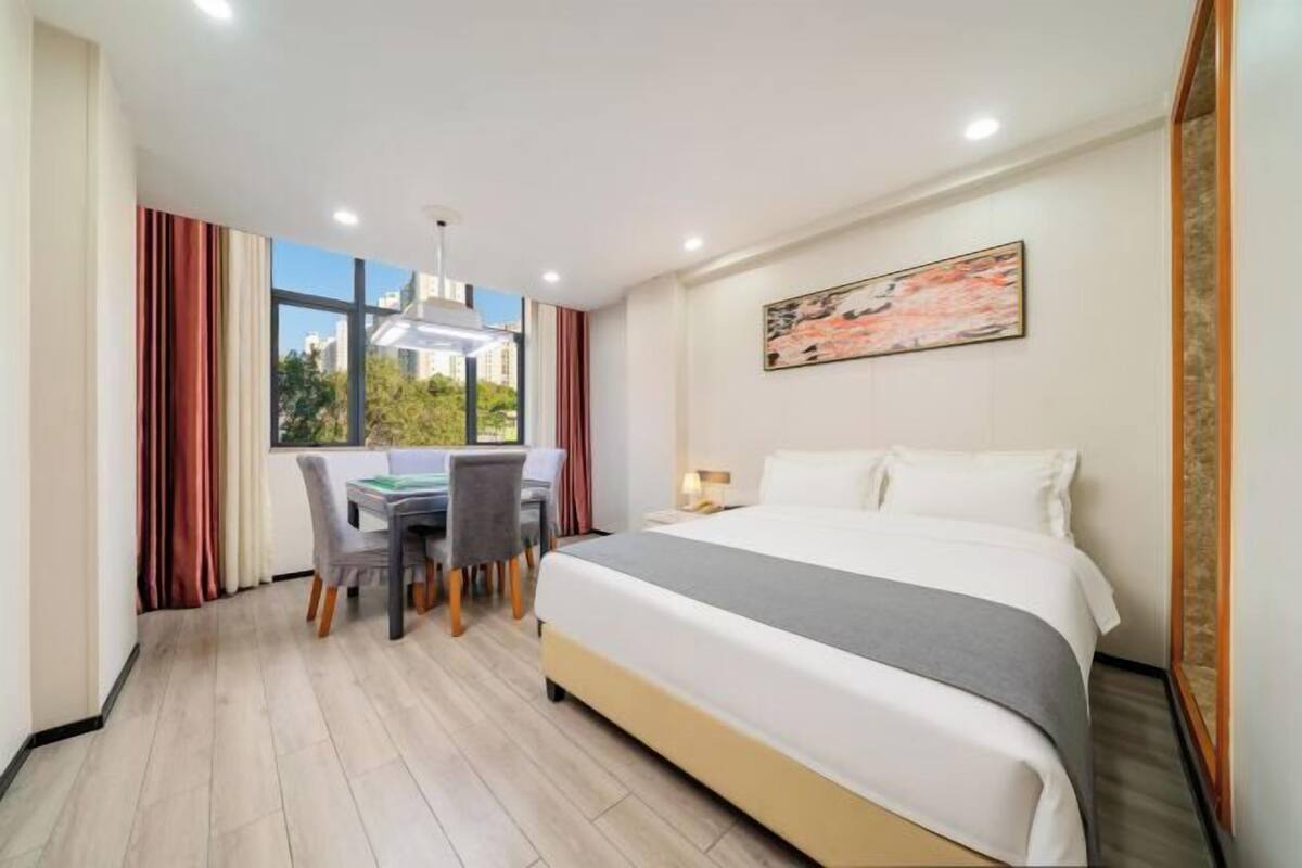 A hotel room with a bed, a desk with chairs, a window with curtains, and a painting on the wall.
