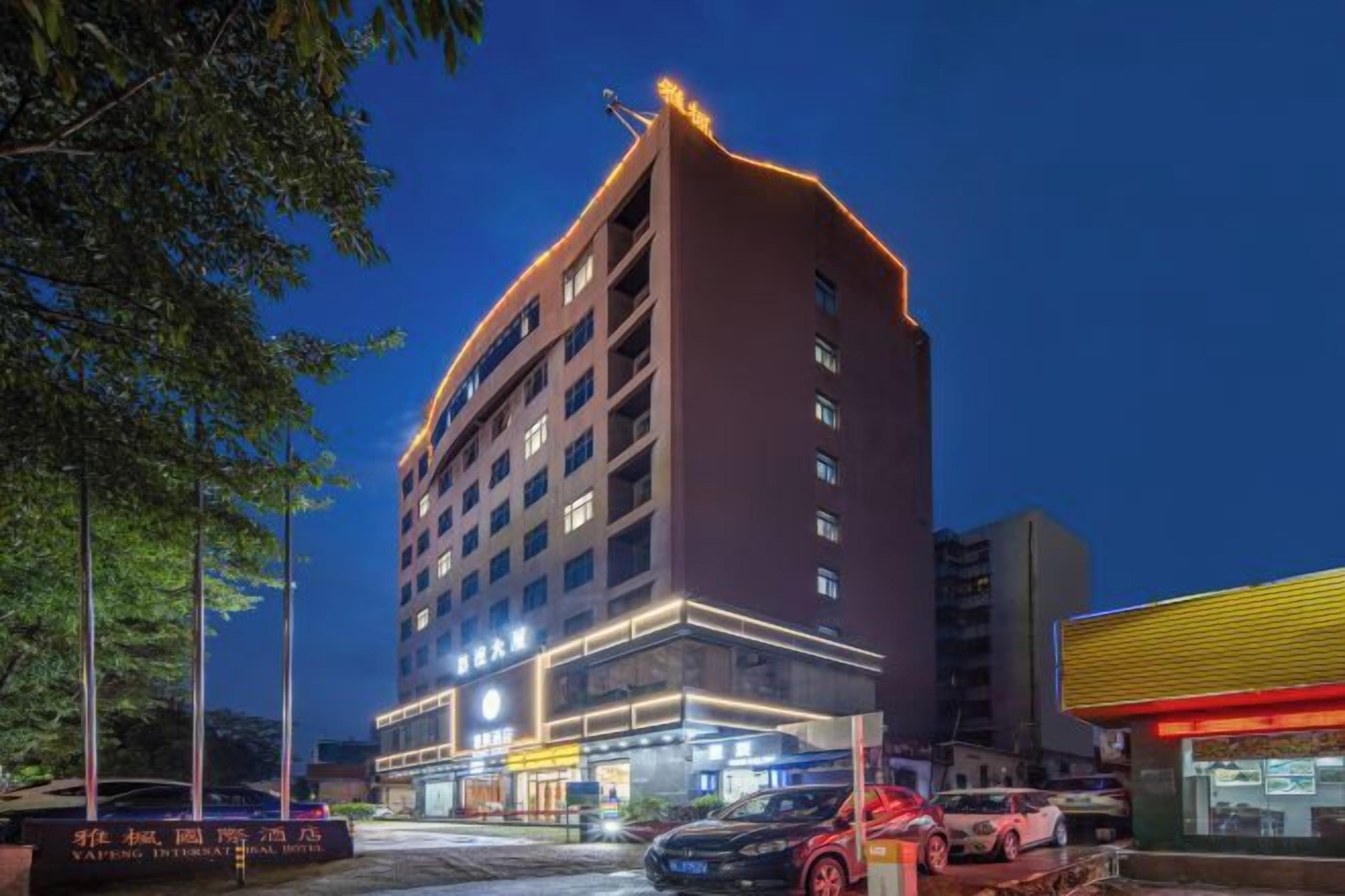Un hotel de varios pisos con una entrada destacada en esquina, un estacionamiento con varios autos y un edificio con toldo amarillo a la derecha.