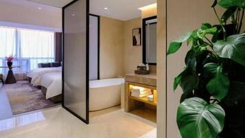 A modern hotel room with a large bed, a separate sitting area, a bathroom with a bathtub, and a large green plant.
