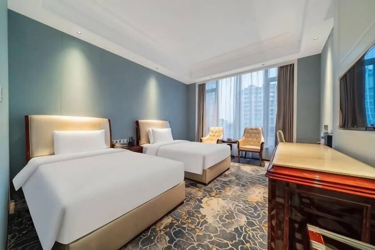 A hotel room with two beds, a desk, a chair, and a large window with a city view.