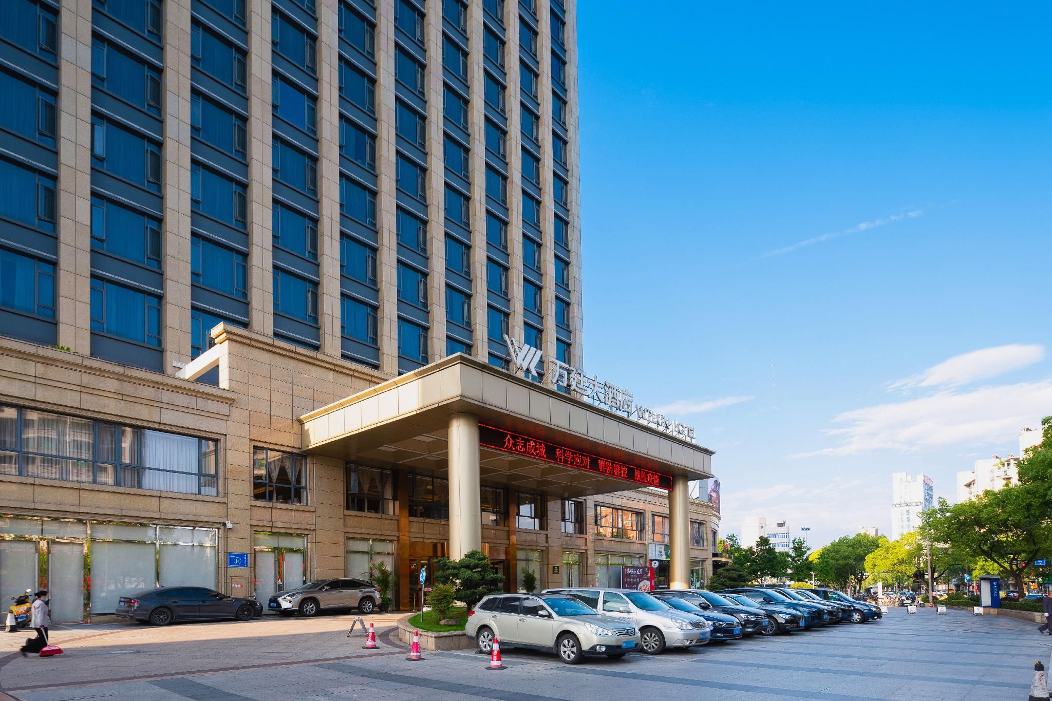A hotel entrance with a modern facade, a covered entrance area, and a parking lot with cars.