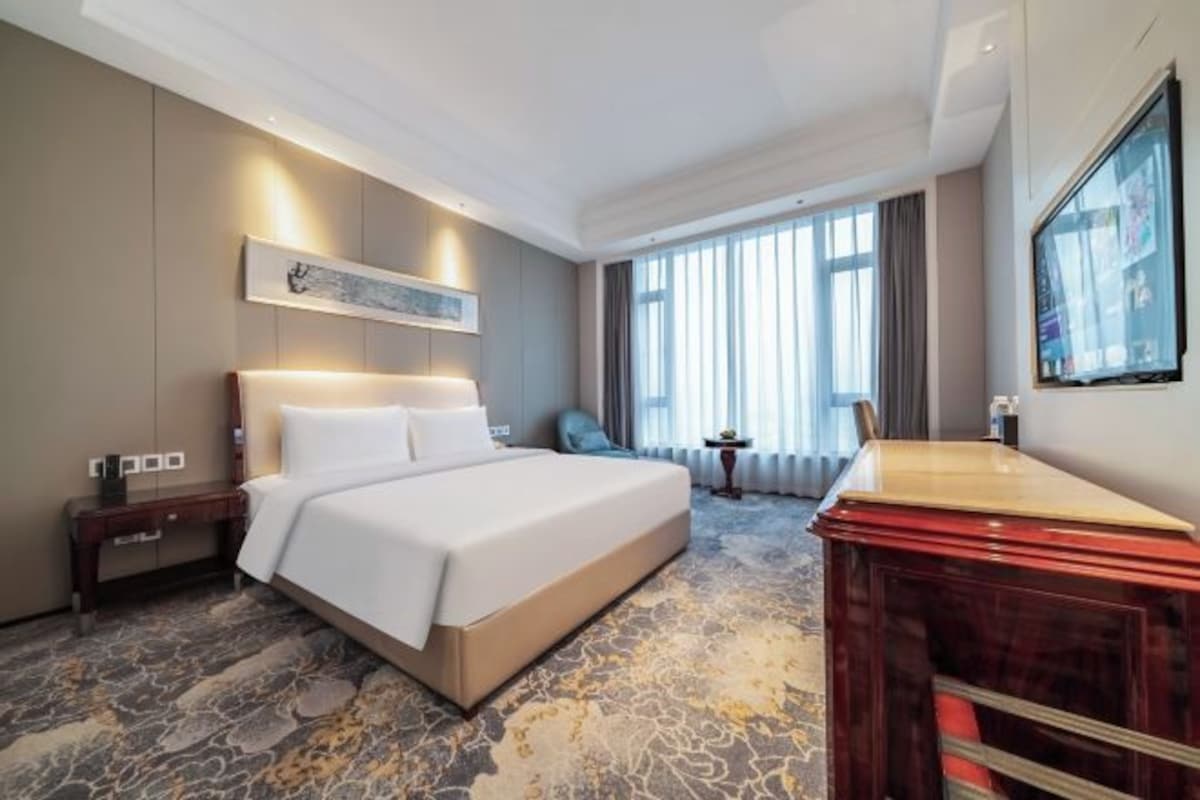 Une chambre d’hôtel avec un grand lit, une commode en bois, une petite table et une grande fenêtre avec des rideaux.