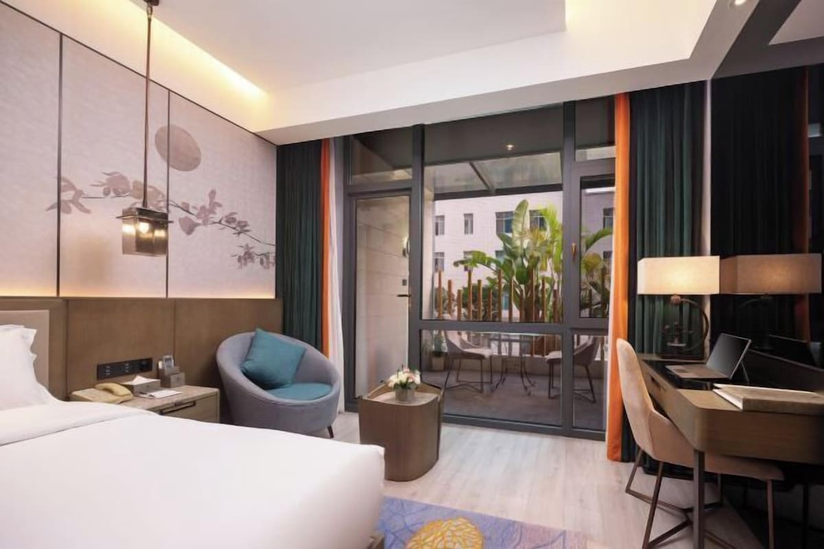 Una habitación de hotel moderna con cama, escritorio, silla, mesita auxiliar pequeña y balcón con vistas a edificios y plantas.