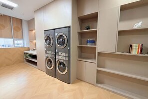 A laundry room with two wall-mounted washing machines, a small seating area, and built-in shelving with books and decorative items.