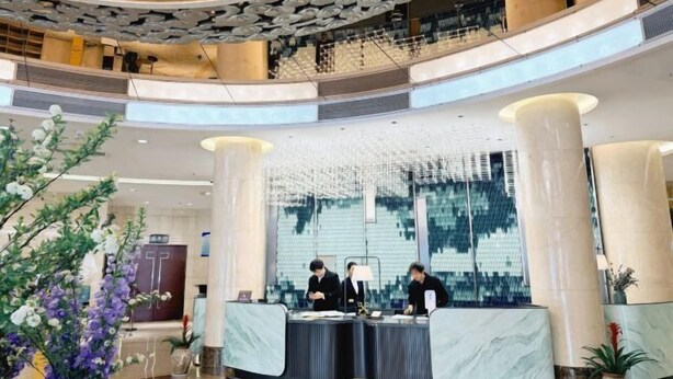 A hotel lobby with a reception desk, two staff members, a decorative ceiling, and a large window. - Zhong Huang Hotel (Kunming)
