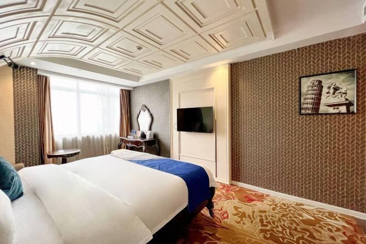 A hotel room with a large bed, a flat-screen TV, a small table, and a patterned carpet.