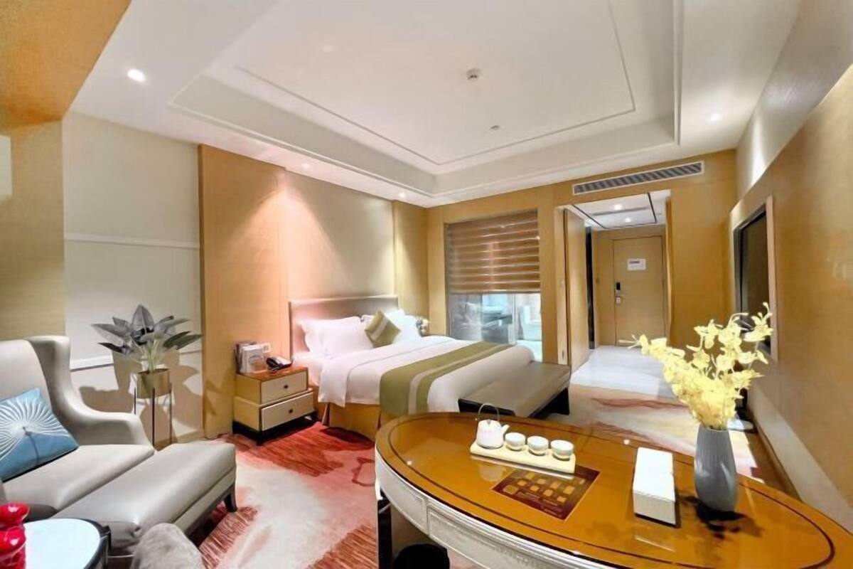 A hotel room with a bed, a desk, a chair, a bedside table, and a vase with flowers.