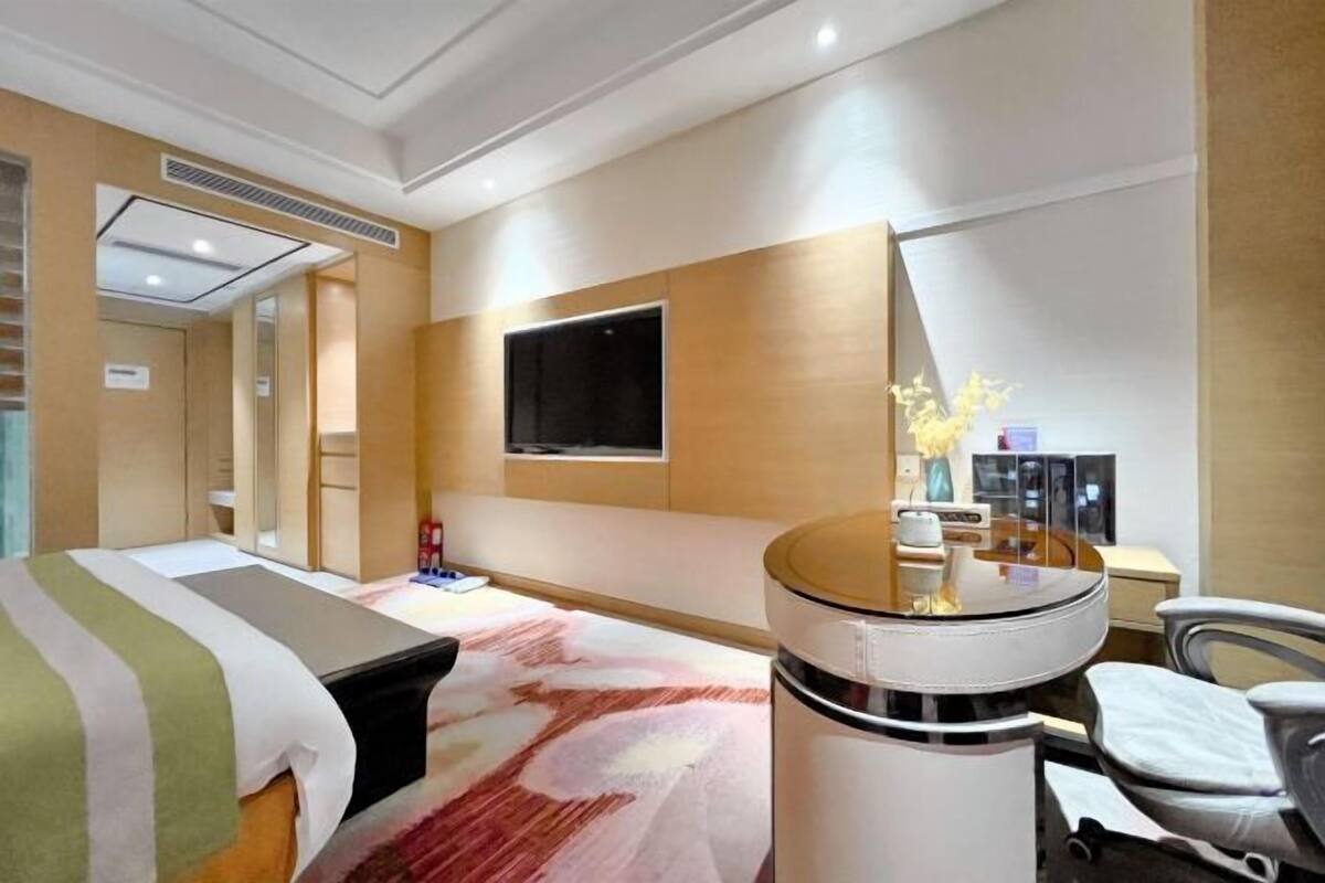 A hotel room with a bed, a desk, a chair, a TV, and a small table with a vase.