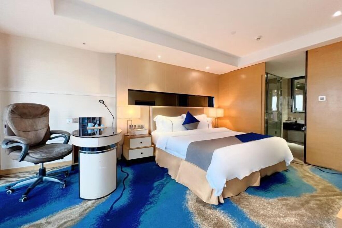A modern hotel room with a bed, desk, chair, and a bathroom visible through a glass door.
