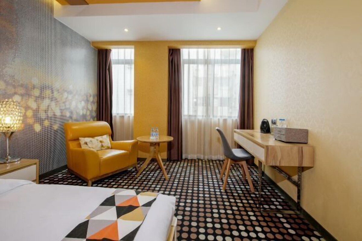 A hotel room with a bed, a desk, a chair, a small table, and a lamp.