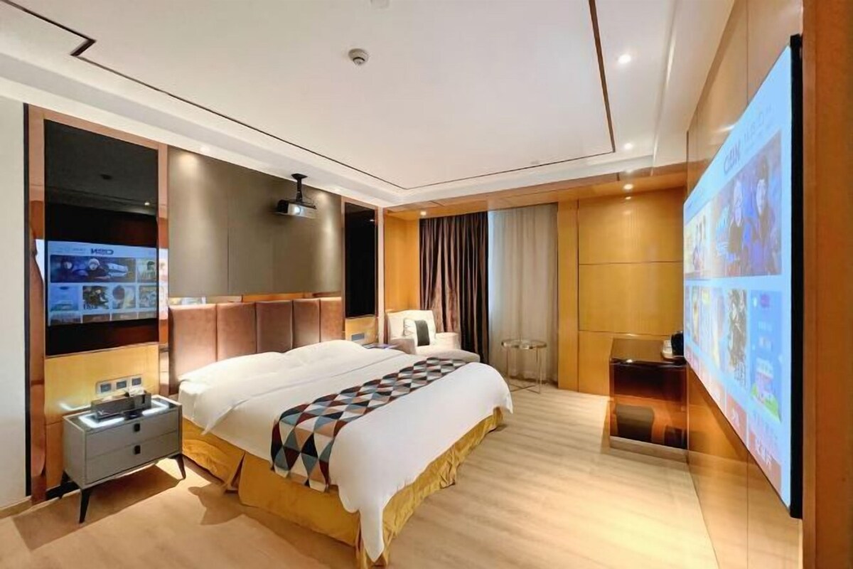 A modern hotel room with a large bed, a flat-screen TV, a bedside table, and a large wall-mounted TV.