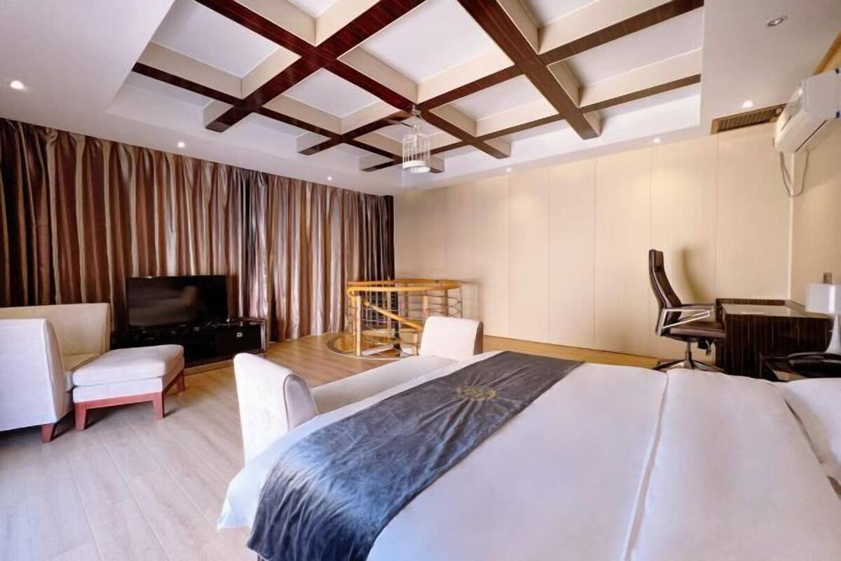 A modern hotel room with a large bed, a desk, a chair, a television, and a ceiling with wooden beams.