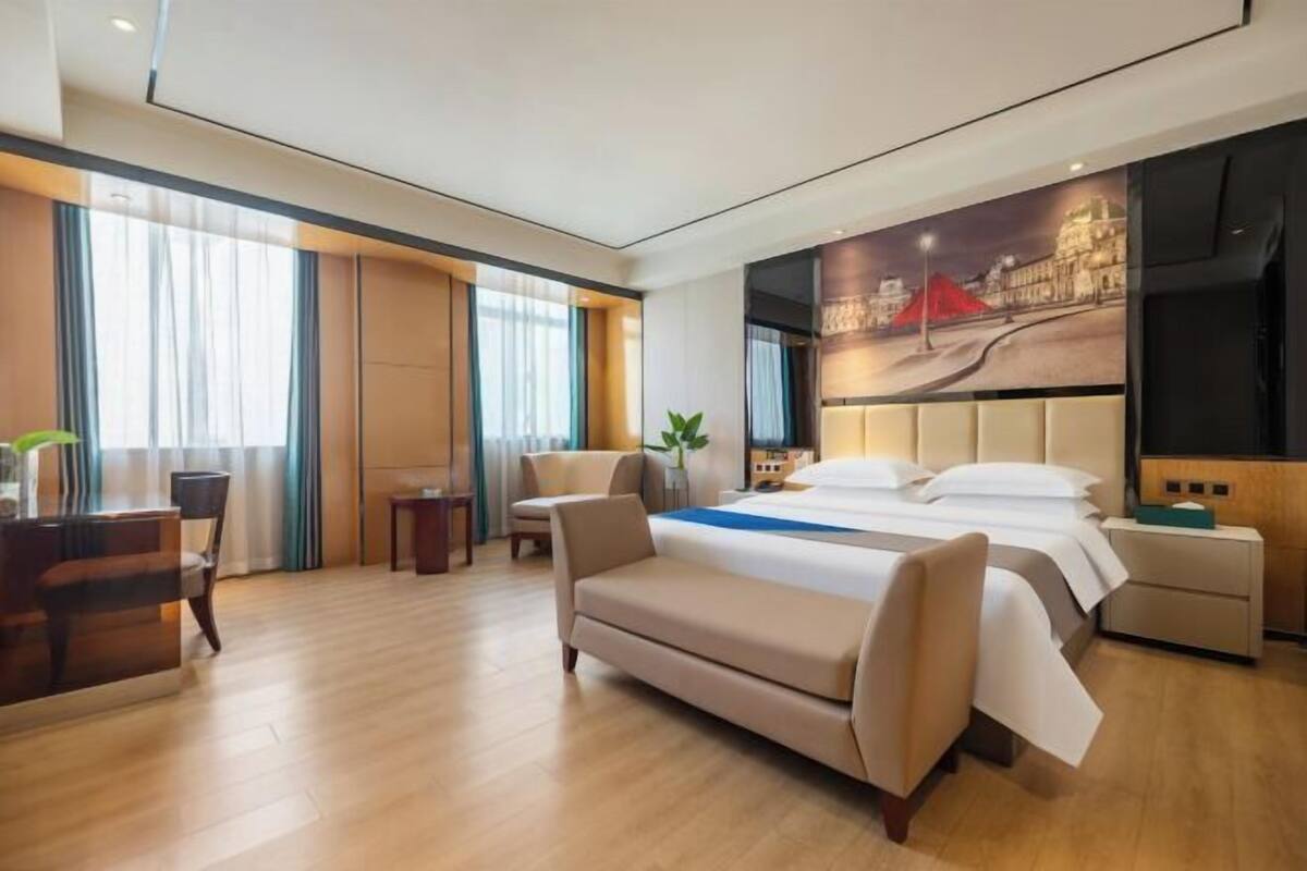 A modern hotel room with a large bed, a desk, a sofa, and a painting on the wall.