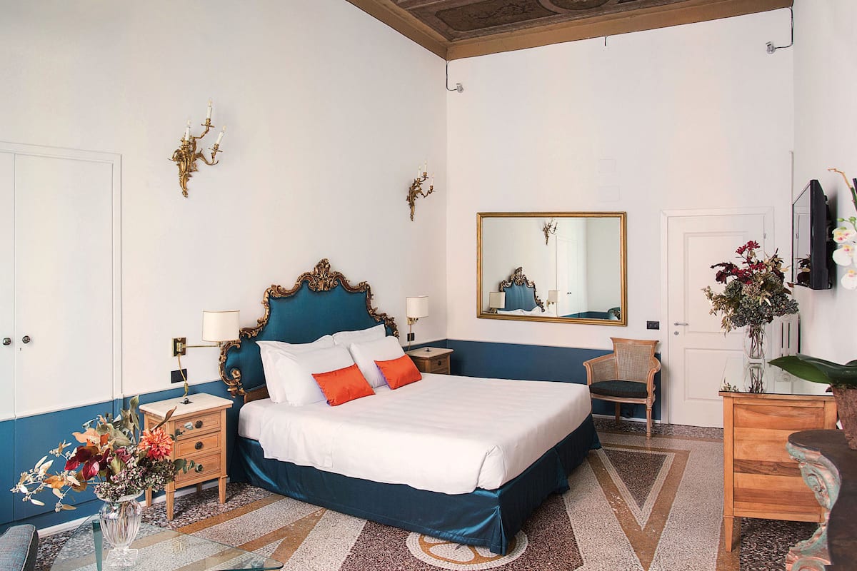 Un dormitorio con una cama grande, mesitas de noche, una silla, un aparador, un espejo y un florero con flores.