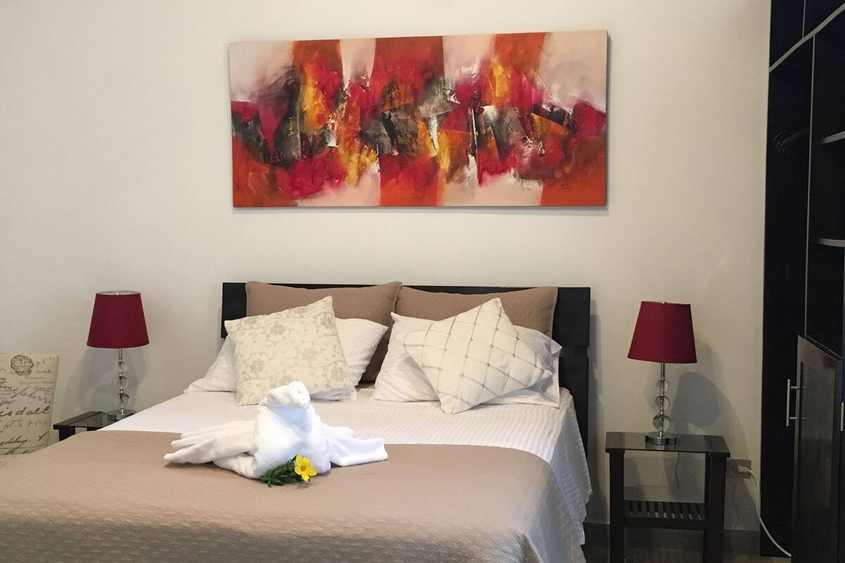 Ein ordentlich bezogenes Bett mit weißer Daunendecke, mehreren Kissen und einem dekorativen weißen Handtuch mit Blumenmuster. Zwei Nachttische mit roten Lampenschirmen, ein weiterer Nachttisch mit Lampe sowie ein großes abstraktes Gemälde über dem Bett.