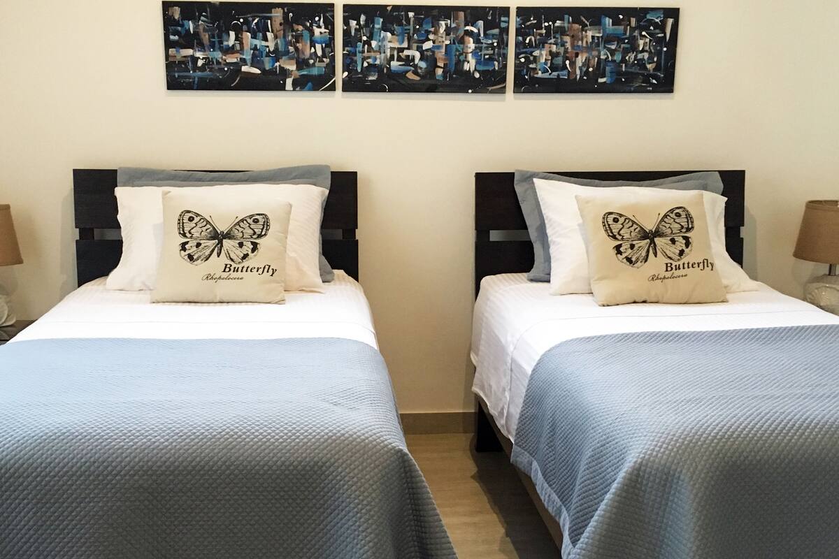 Ein Hotelzimmer mit zwei Betten, jedes mit einem Kissen mit einem Schmetterlingsmotiv und dem Text "Butterfly Rhopalocera". Darüber an der Wand befinden sich drei abstrakte Gemälde.