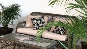 A wicker sofa with beige cushions and black floral pillows, a glass coffee table, and green plants. - Las Perlitas Boutique Hotel (Sámara)