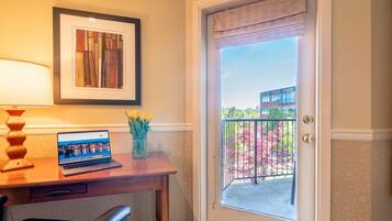 A room with a desk, chair, laptop, framed artwork, and a door leading to a balcony with a view of buildings and trees.
