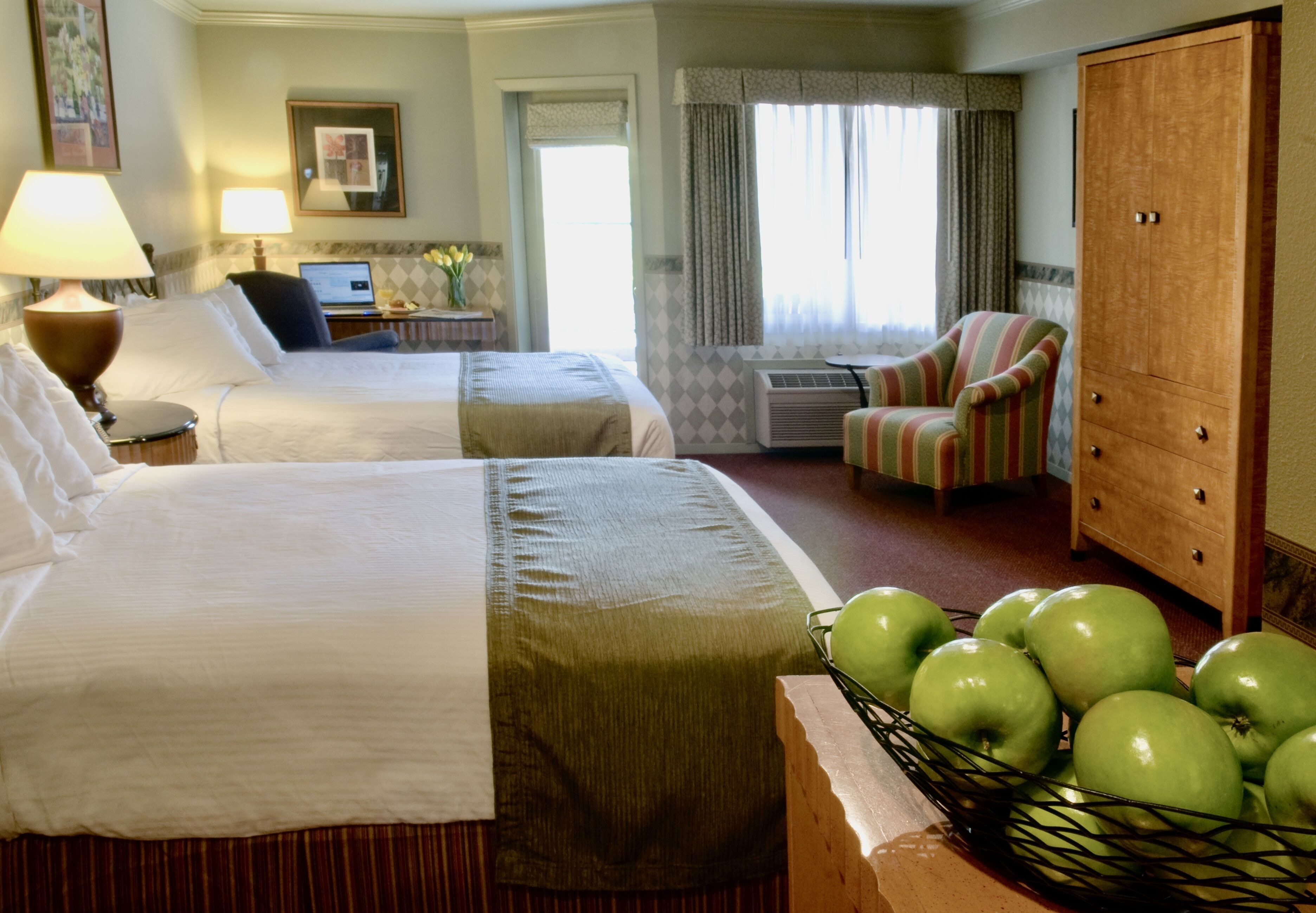 A hotel room with two beds, a desk with a laptop, a chair, a wardrobe, and a basket of green apples.