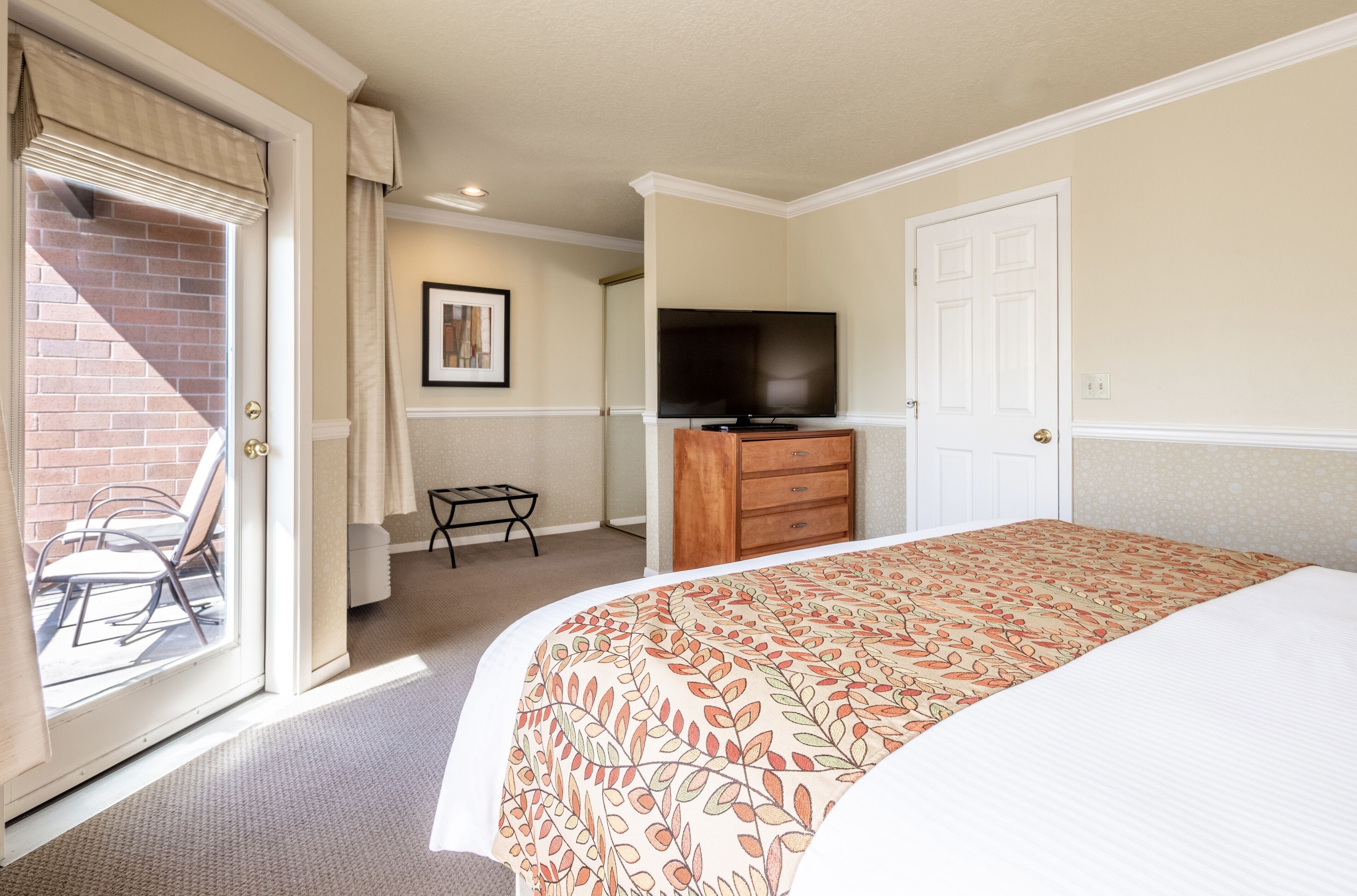 Habitación de hotel con una cama, televisión, cómoda de madera, una mesita y una silla cerca de un ventanal corredera.