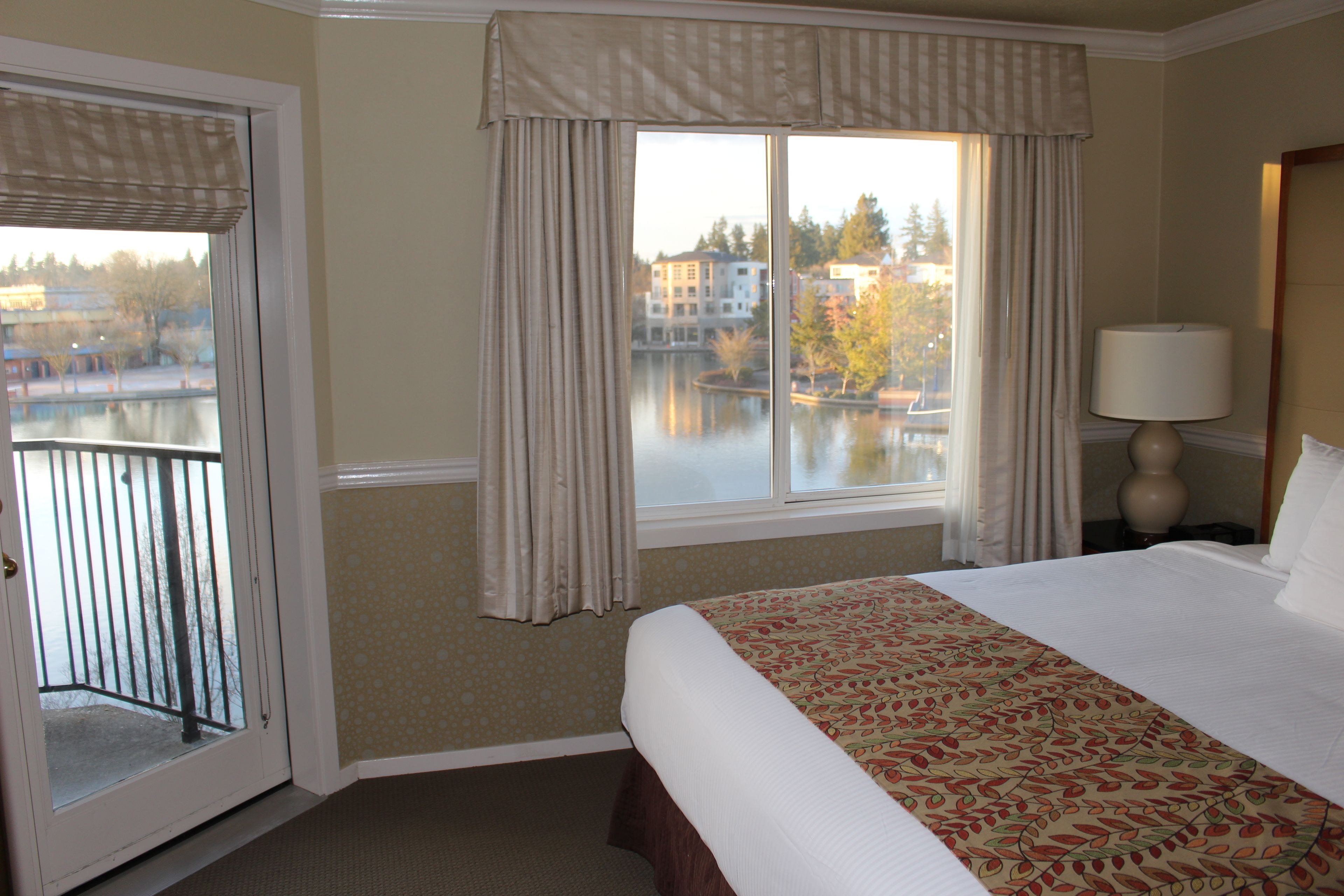 A hotel room with a large bed, a window with curtains, a view of a lake, and a balcony.