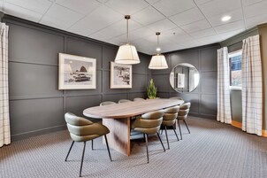 A conference room with a long table, six chairs, two pendant lights, framed pictures on the wall, a round mirror, and a window with curtains.