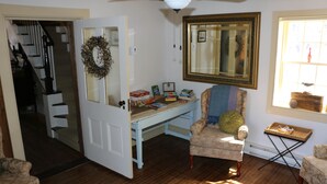 A cozy living room with a wooden floor, a desk with books, a chair, a mirror, and a staircase leading to another room.