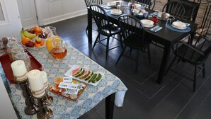 A dining area with a table set for four, featuring a patterned tablecloth, black chairs, and various food items including fruits and a pitcher.