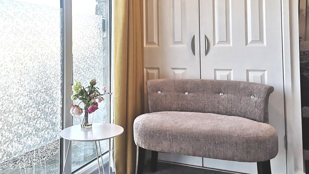 Una habitación con un ventanal grande, una mesita redonda con un florero, un banco tapizado gris y un armario blanco de dos puertas.