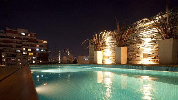 Una piscina nocturna con agua cristalina, rodeada de muros de piedra y plantas en macetas, con edificios al fondo.