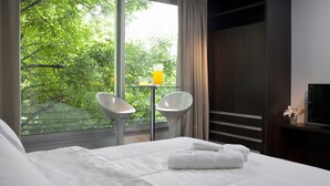 Habitación de hotel con cama, dos sillas blancas, una mesita con un vaso de jugo de naranja y un televisor sobre un mueble de madera.