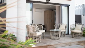 Un área de estar al aire libre con sillas de mimbre y una mesa de vidrio, una pérgola de madera y una puerta corrediza que da a un espacio interior.