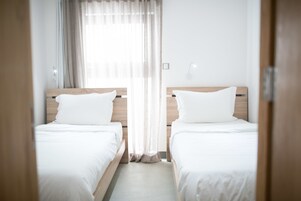 Habitación de hotel con dos camas individuales, cabecero de madera, ventana con cortinas translúcidas y un aplique de pared.