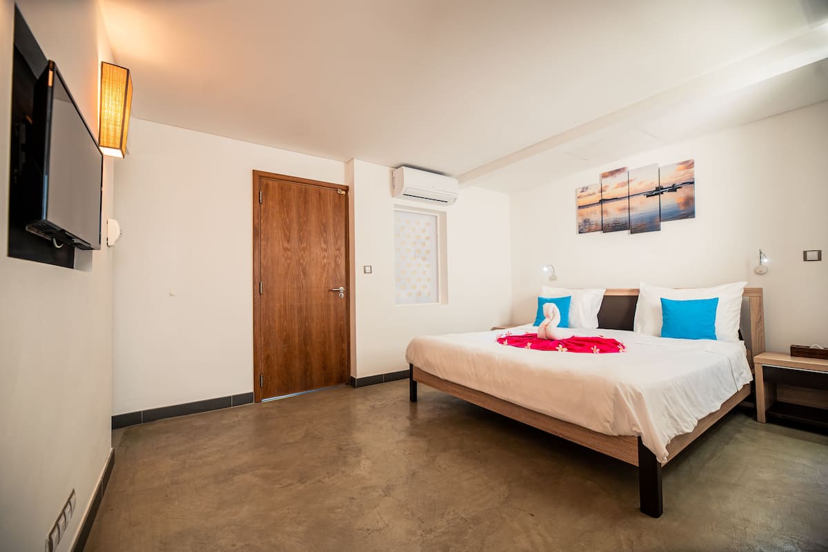 A hotel room with a bed, a TV mounted on the wall, an air conditioning unit, and a painting above the bed.