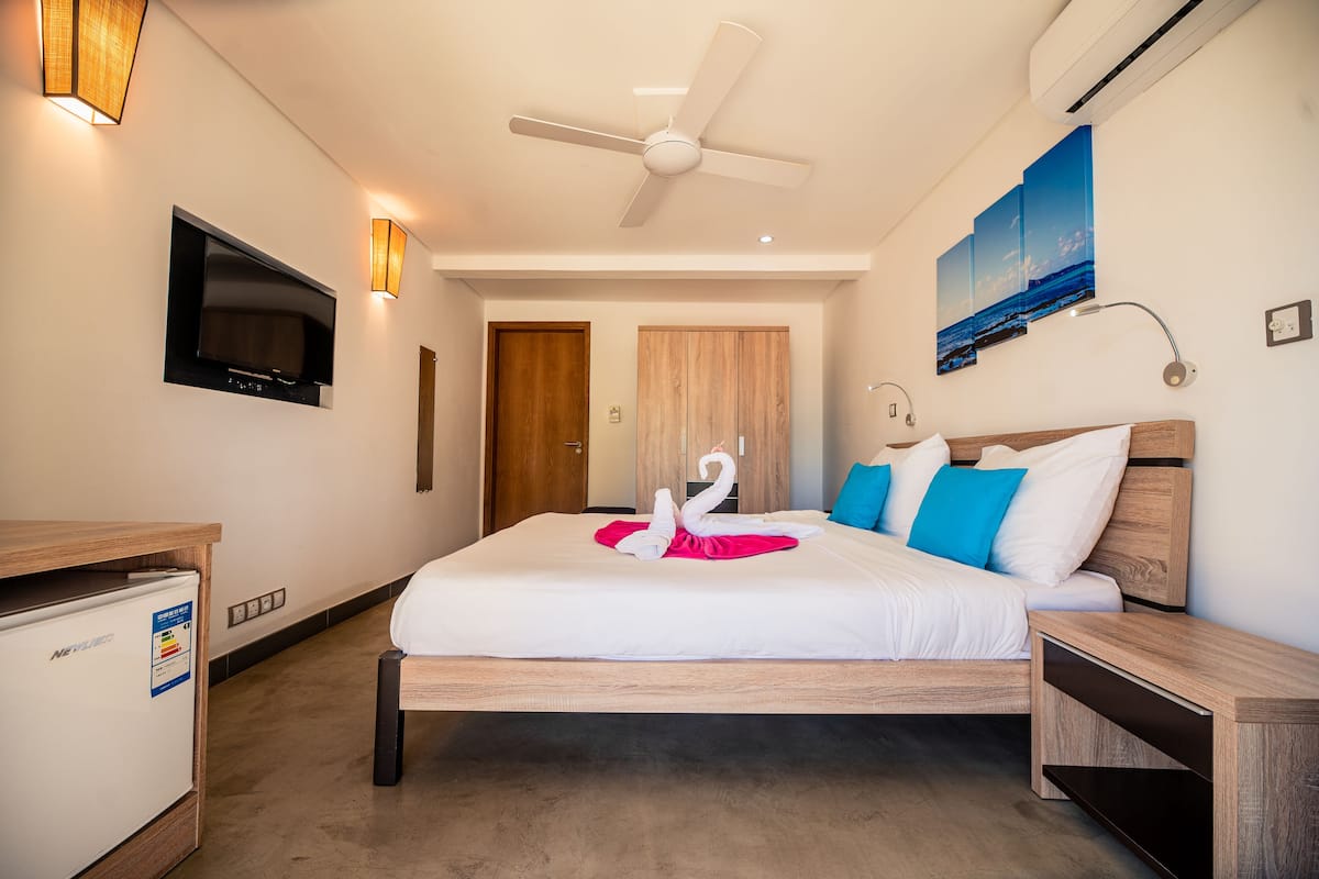A hotel room with a bed, a ceiling fan, a TV, a wall-mounted lamp, and a painting.