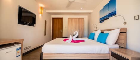 A hotel room with a bed, a ceiling fan, a TV, a wall-mounted lamp, and a painting.