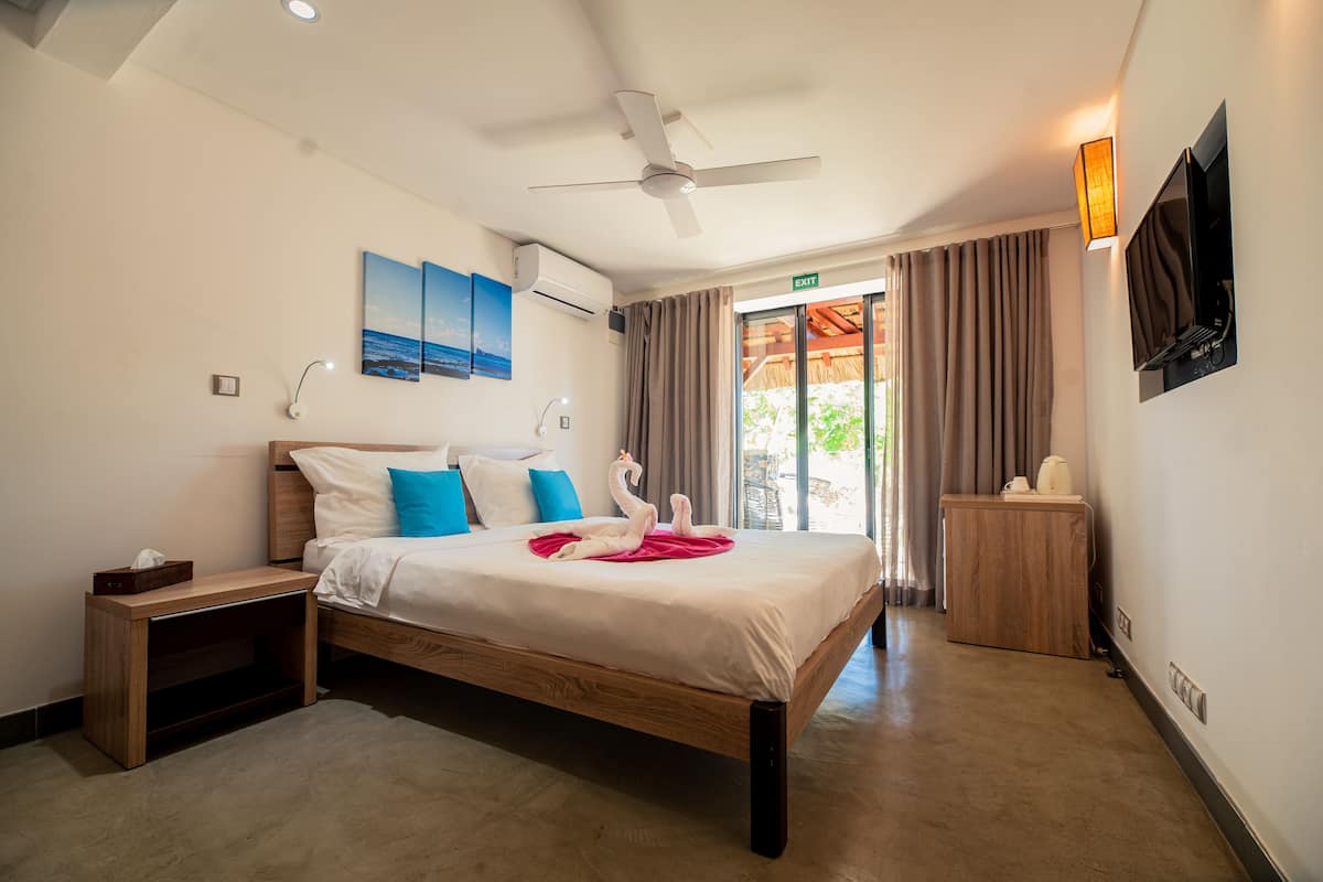A hotel room with a bed, a nightstand, a wooden side table, a ceiling fan, a TV, and a window with curtains.