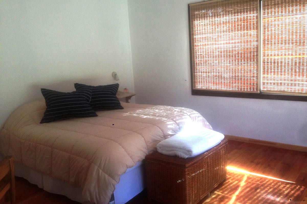 A bedroom with a bed, pillows, a nightstand, a window with blinds, and a wooden floor.